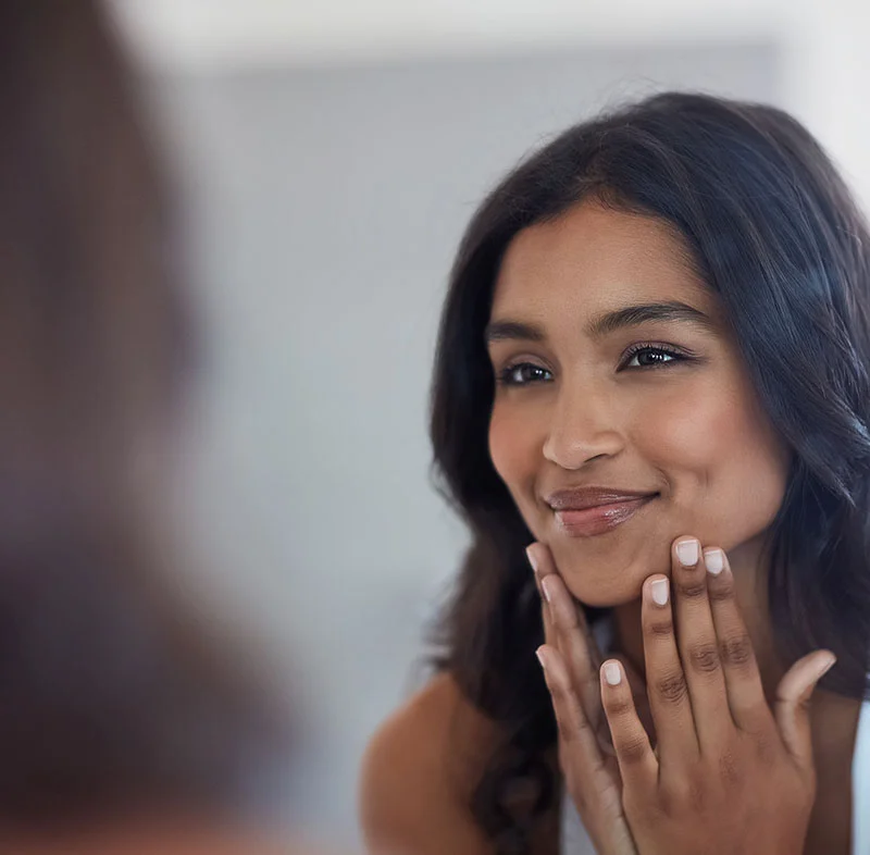 Smiling woman touching her chin while admiring her reflection in a mirror, with a soft and natural expression - Acne Scar Treatment in North Carolina