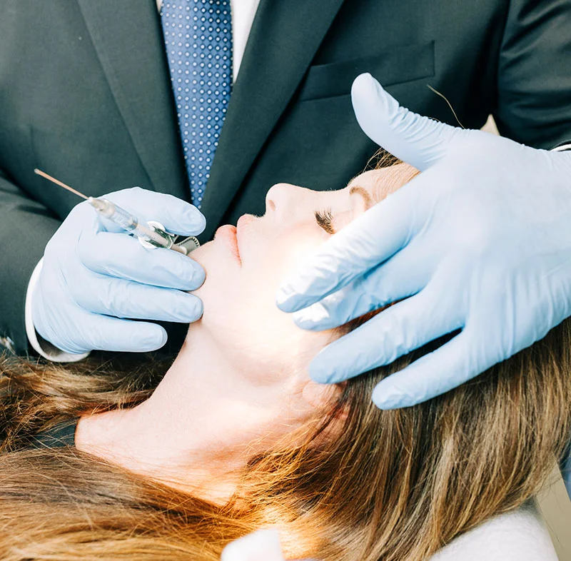 Close-up of a cosmetic procedure where a doctor in a suit administers an injection to a woman's chin, wearing blue gloves - Cheek Fillers in North Carolina