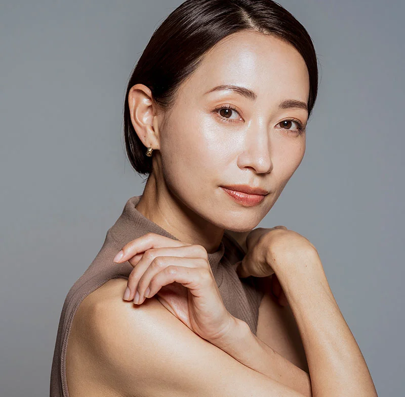 A close-up of an elegant Asian woman with smooth skin, short hair, and a calm expression, wearing a taupe-colored top, gently holding her arms near her chest in a graceful pose against a neutral background - Chemical Peels in North Carolina