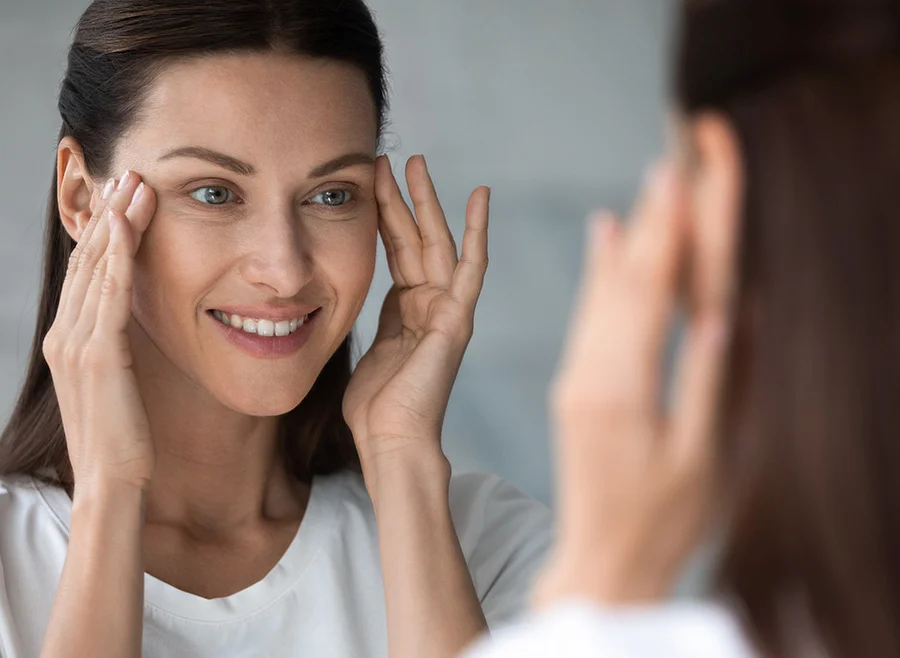 A woman with dark hair gently touches her face with both hands while looking into a mirror, smiling softly, as she examines her skin - Chemical Peels in North Carolina