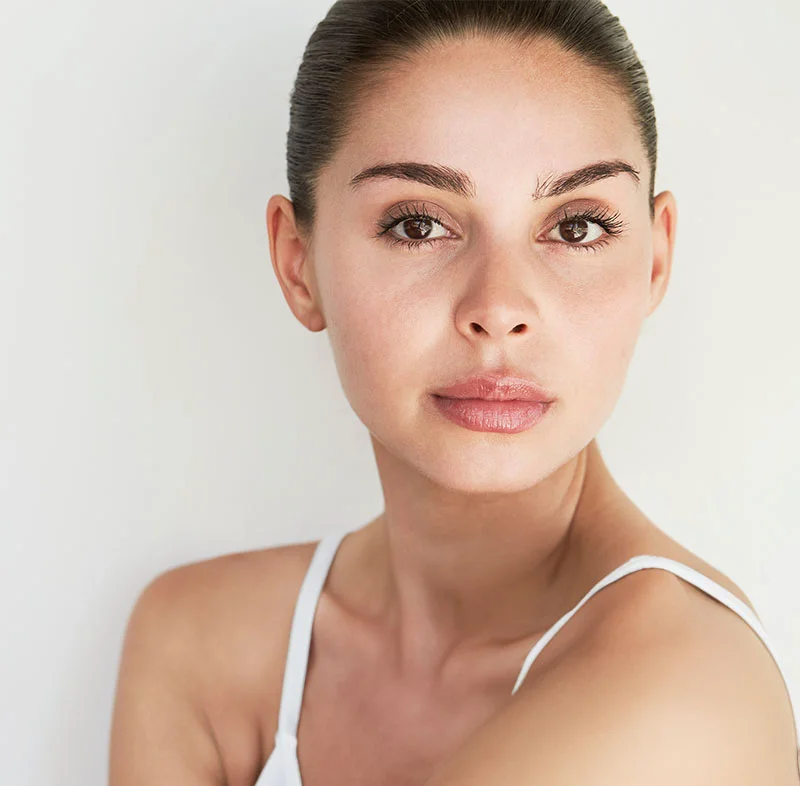 A close-up of a woman with clear skin and natural makeup, wearing a white tank top, looking directly into the camera with a relaxed and soft expression - Dermal Fillers in North Carolina