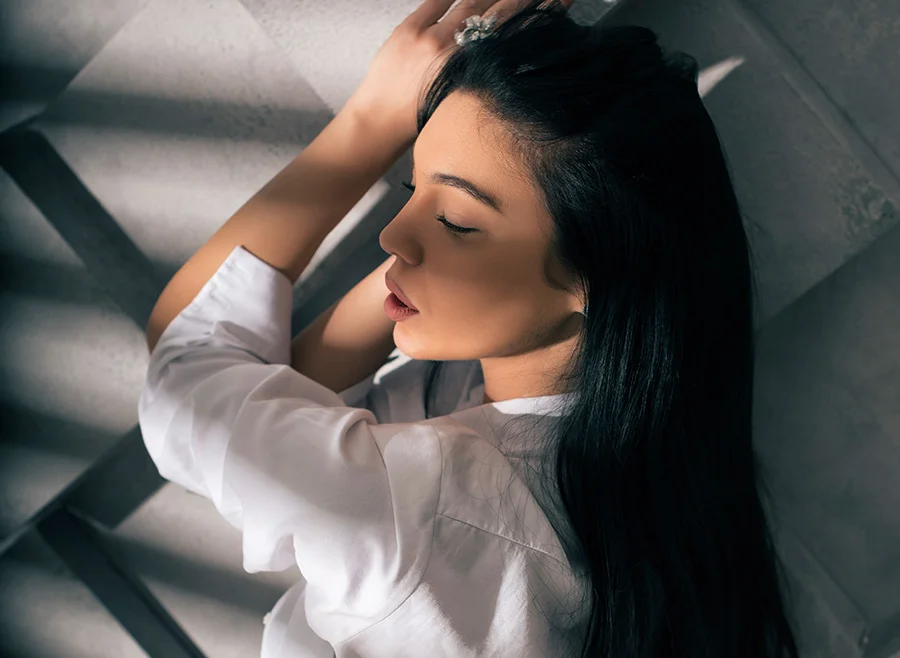 A woman with long dark hair and closed eyes leans against a textured wall, wearing a white shirt, and basking in soft, shadowy light - Dermal Fillers in North Carolina