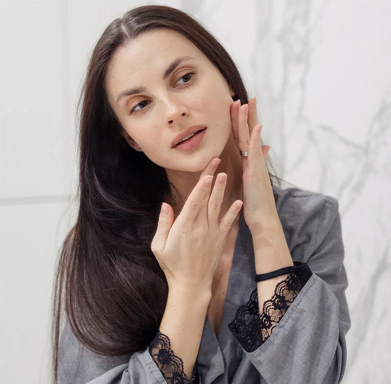 A woman with long dark hair, wearing a gray robe, gently touches her face with one hand, examining her skin with a thoughtful expression - Dermaplaning in North Carolina