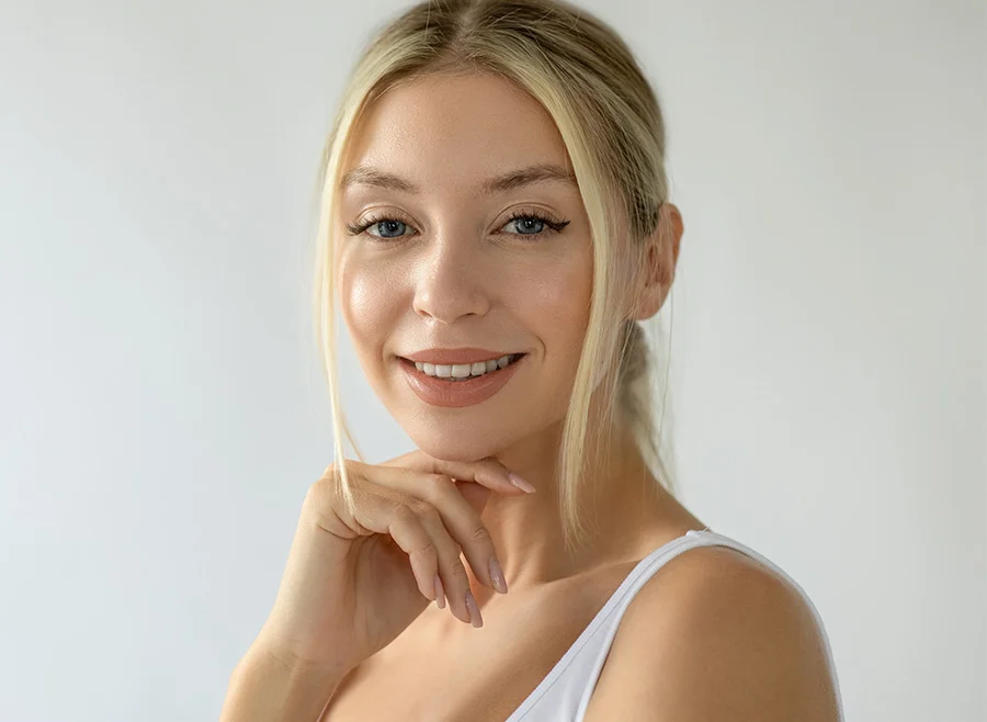 A blonde woman with a soft smile, wearing a white tank top, poses with her hand gently resting under her chin, exuding a fresh and natural beauty against a light background - Dermaplaning in North Carolina