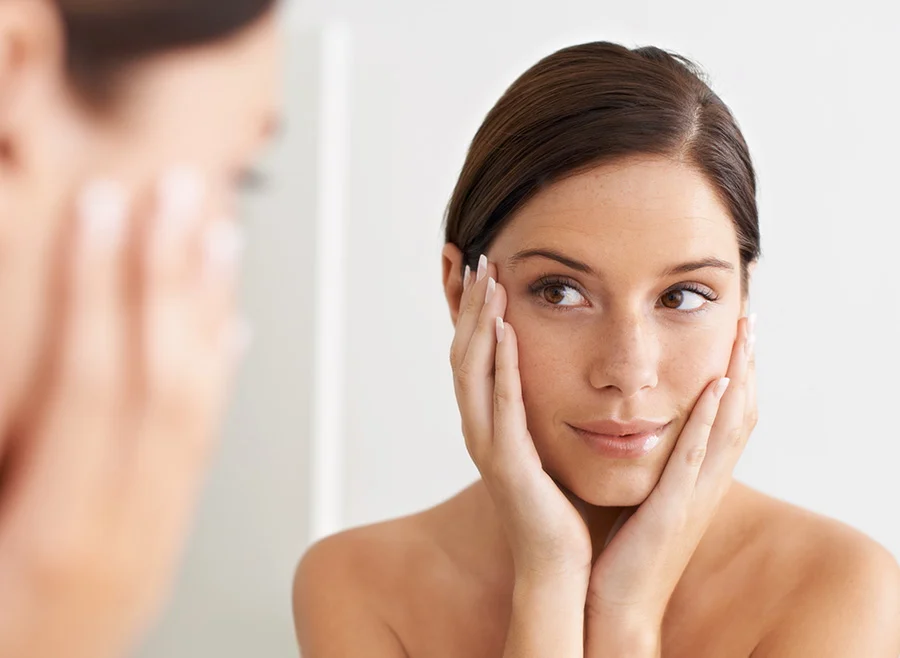 Young woman with smooth skin gently touching her face, looking at her reflection in the mirror - Diamond Glow Facial in North Carolina