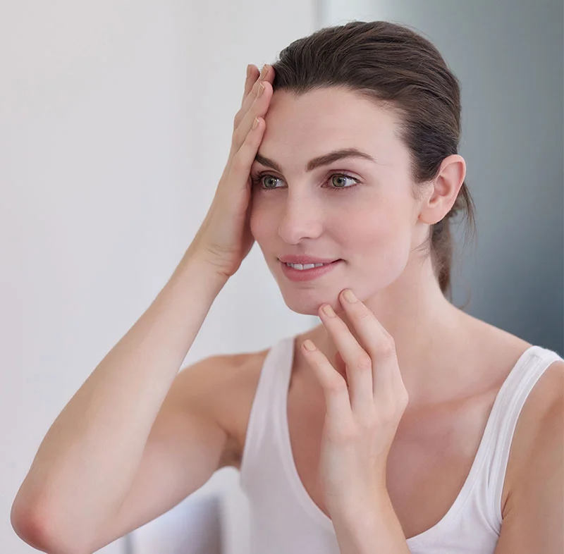 Woman with her hand on her forehead and chin, smiling softly as she examines her skin in the mirror - Face Tight in North Carolina