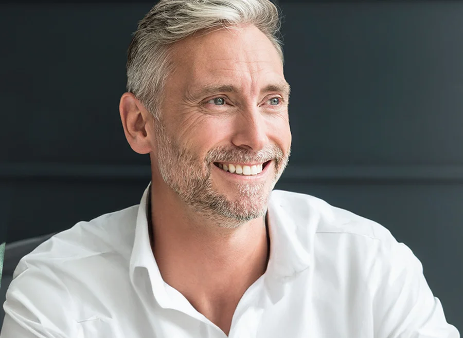 A smiling mature man with salt-and-pepper hair, wearing a white shirt, looking off to the side against a dark background - Hair Loss Specialist in North Carolina