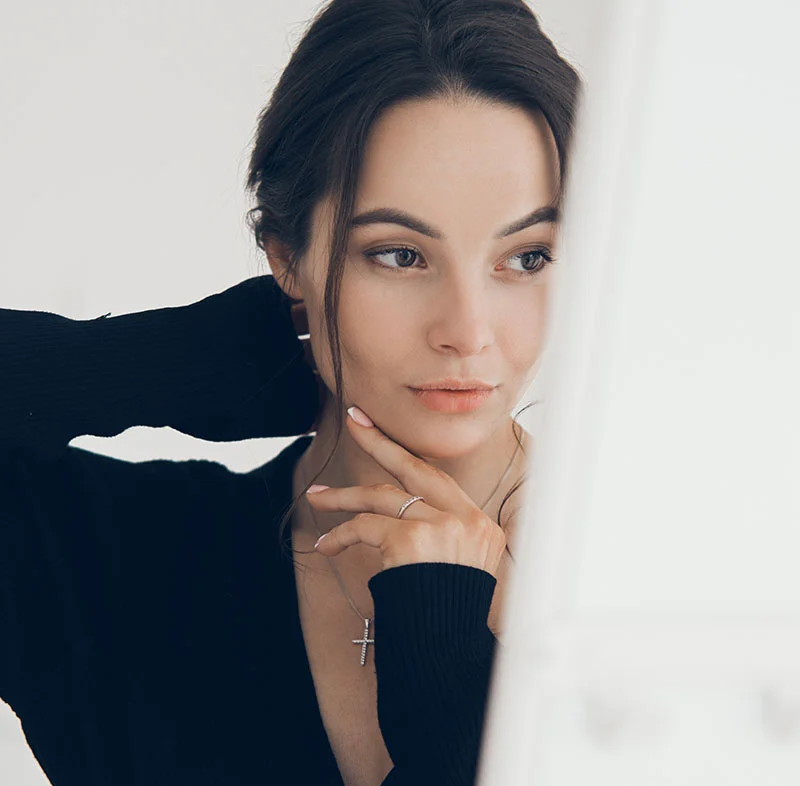 A thoughtful young woman with dark hair tied back, wearing a black sweater, gently touching her neck while looking in the mirror - Hydrafacial in North Carolina