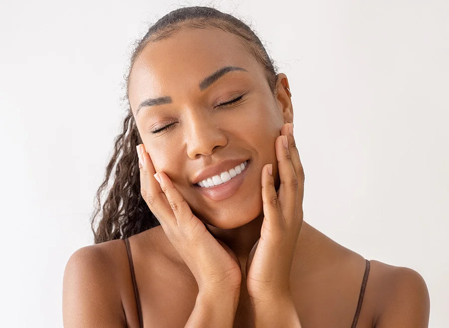 A woman with glowing skin and closed eyes, smiling and touching her face with both hands, exuding a sense of calm and happiness - Hydrafacial in North Carolina