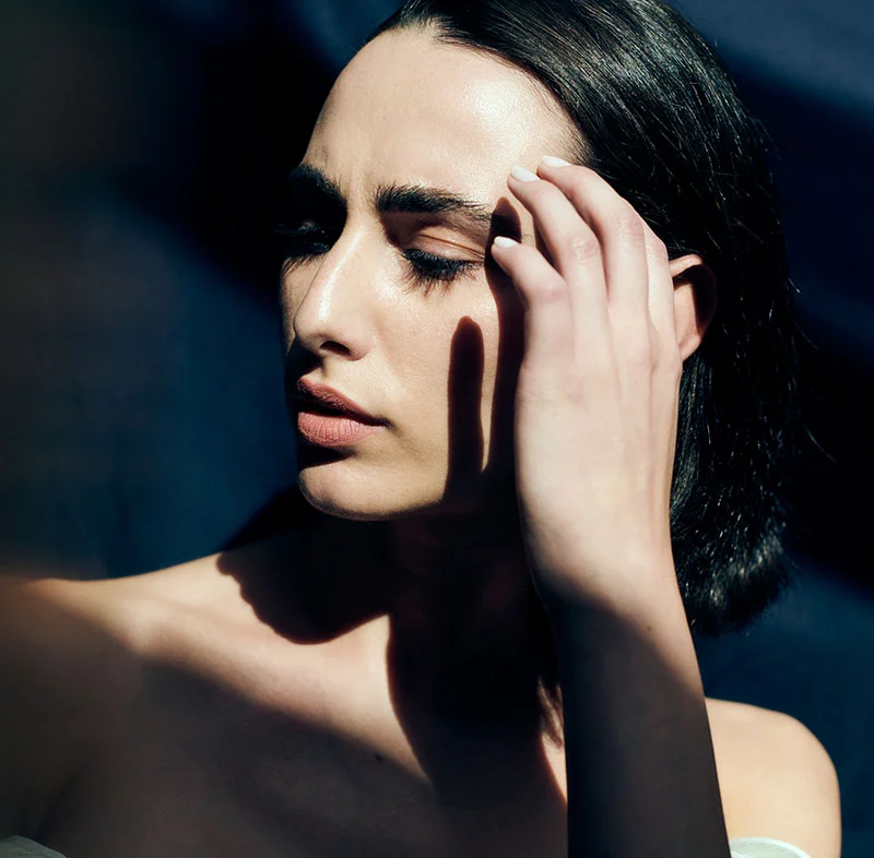 A woman with short dark hair, in a dramatic light and shadow, closes her eyes and gently touches her forehead with one hand - Laser Skin Resurfacing in North Carolina