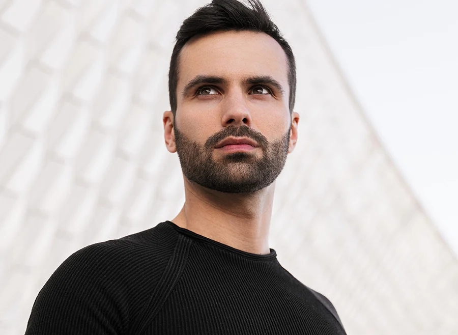 A man with a neatly trimmed beard and serious expression, standing outdoors in front of a modern, white architectural background, dressed in a black shirt - Male Pattern Baldness in North Carolina