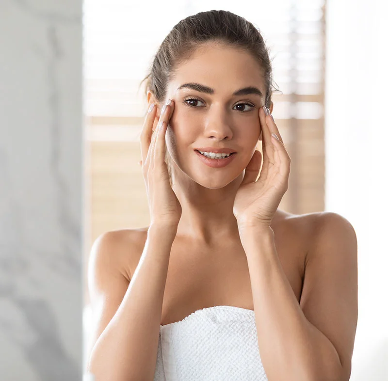 A woman with a towel wrapped around her body gently touching her face, looking into the mirror with a relaxed expression in a well-lit bathroom - Morpheus 8 RF Microneedling in North Carolina