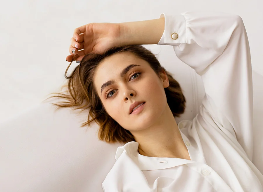 A young woman reclining on a white couch, dressed in a white shirt, with her hand resting on her forehead, gazing thoughtfully towards the camera - Morpheus 8 RF Microneedling in North Carolina