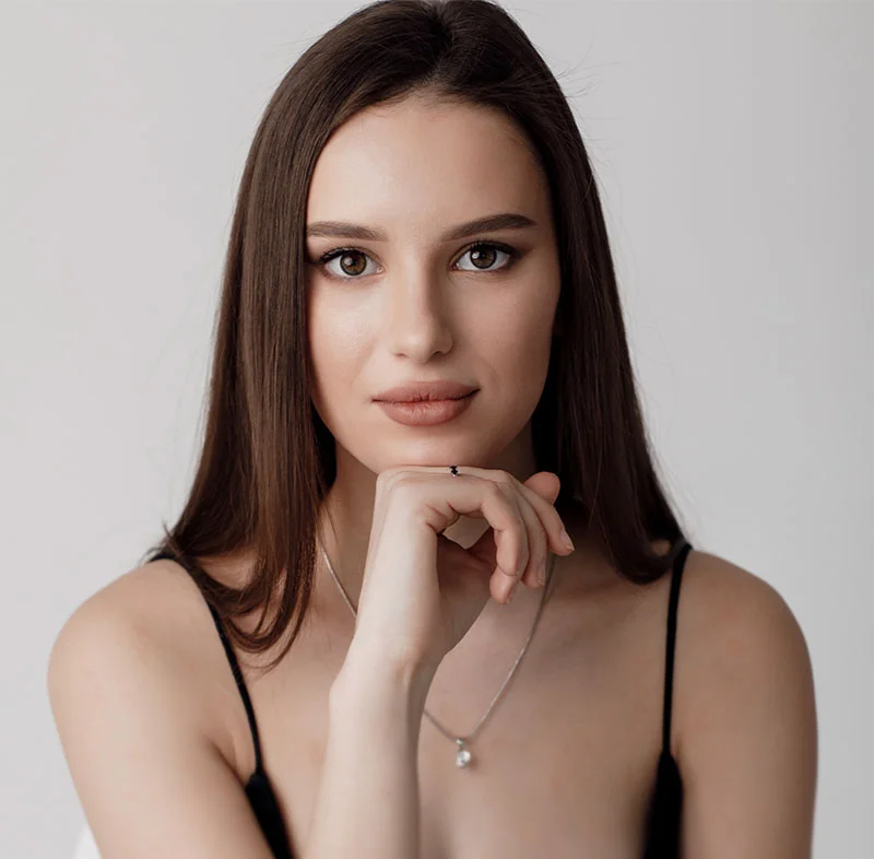 A young woman with long brown hair, wearing a black camisole, resting her chin on her hand while looking directly into the camera, against a light gray background - Permanent Makeup in North Carolina