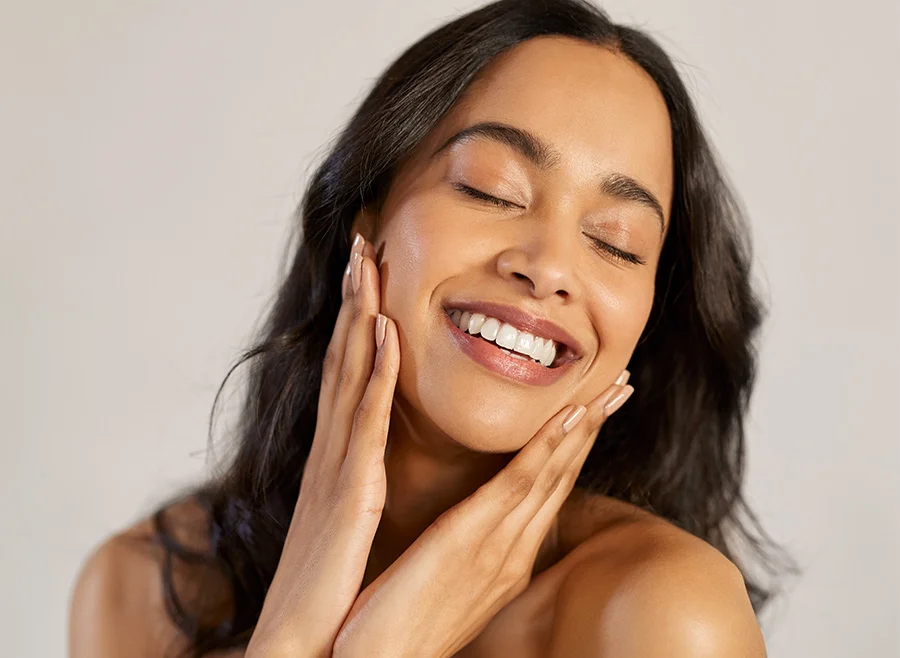 A woman with dark hair and glowing skin, smiling with her eyes closed, gently touching her face with both hands in a moment of relaxation and joy - Permanent Makeup in North Carolina