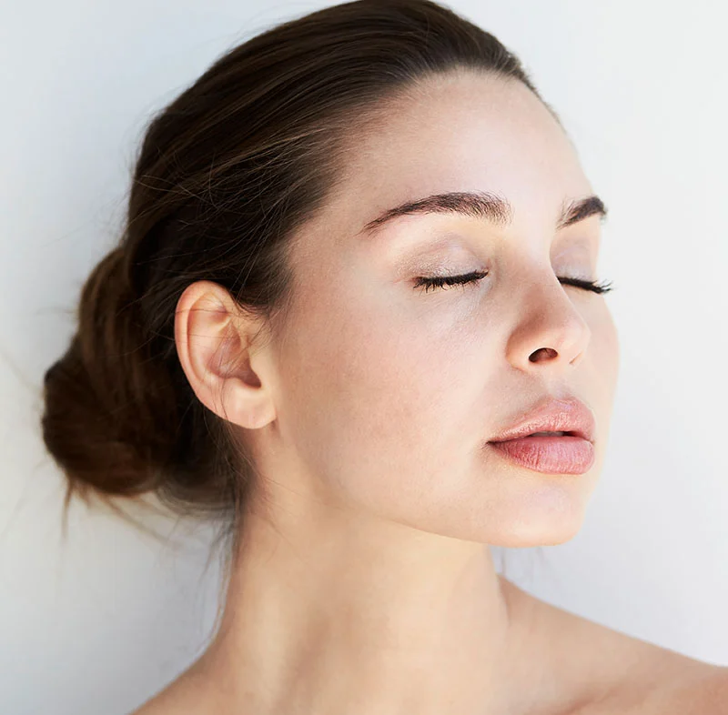 A woman with her eyes closed, in a serene and relaxed pose, her hair tied back in a loose bun, with a neutral expression on her face - PRP for Hair Restoration in North Carolina