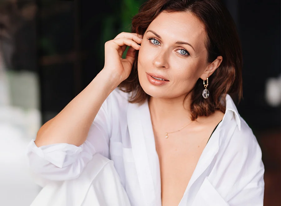 A woman with shoulder-length brown hair and blue eyes, wearing a white blouse, sitting comfortably and resting her head on her hand, smiling softly - PRP Vaginal Rejuvenation in North Carolina