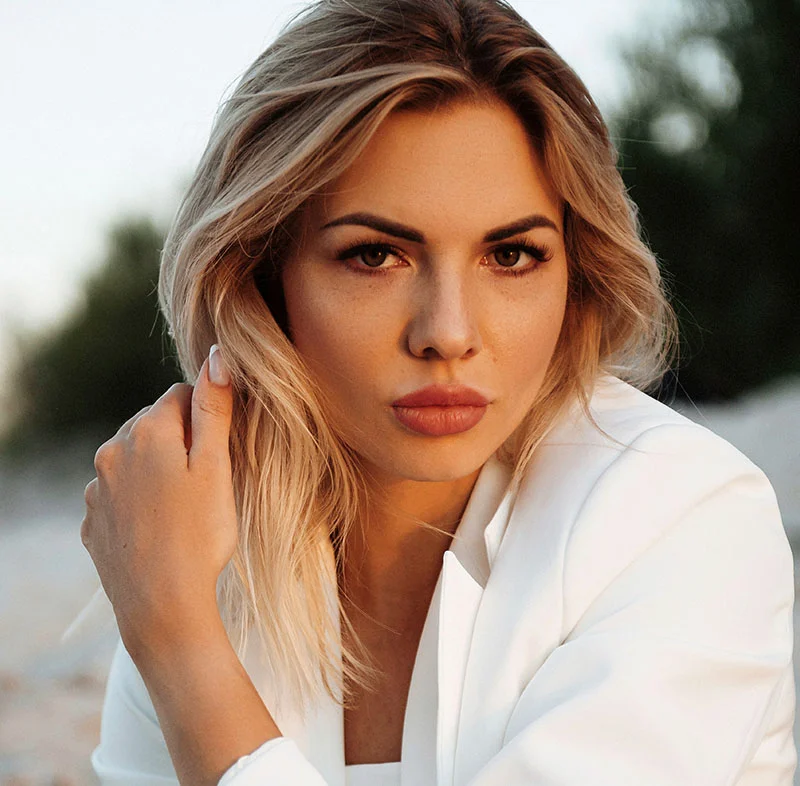 A blonde woman in an outdoor setting, wearing a white jacket, looking intently at the camera with a serious and focused expression - Sculptra Bio Simulator in North Carolina